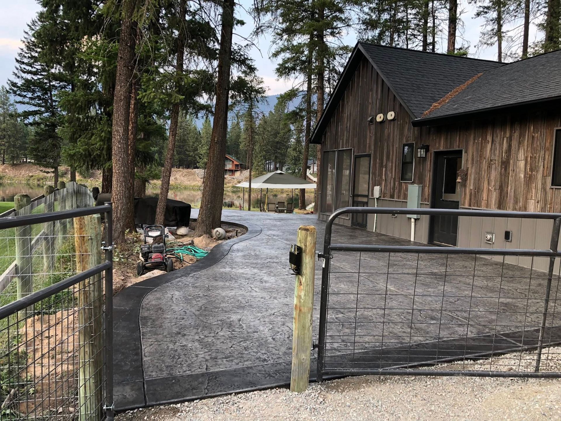 Gravel driveway gate beside a rustic cabin in a wooded area, with pine trees and a parked car in the background