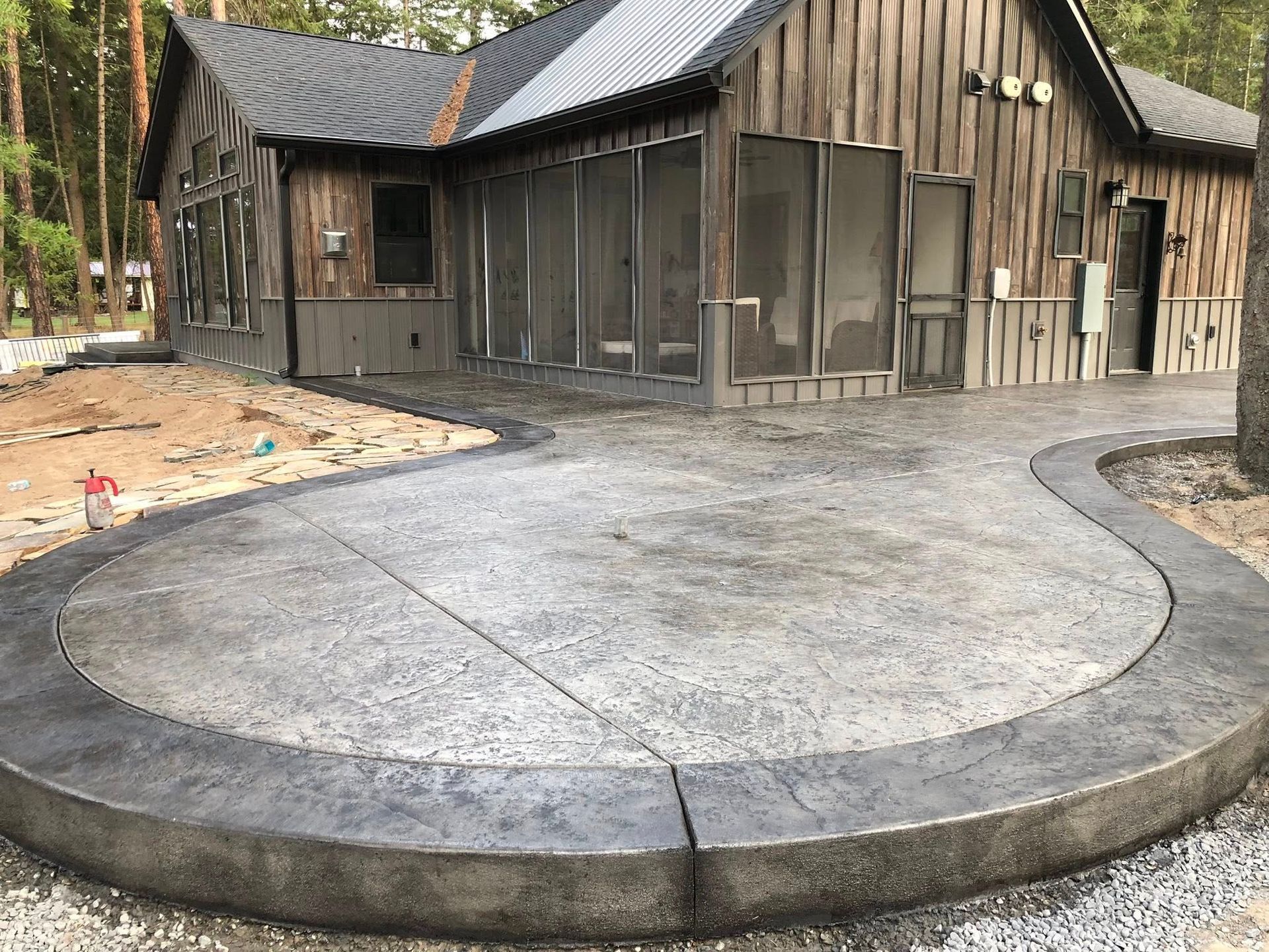 Curved concrete patio in front of a rustic wooden cabin under construction, surrounded by dirt and trees