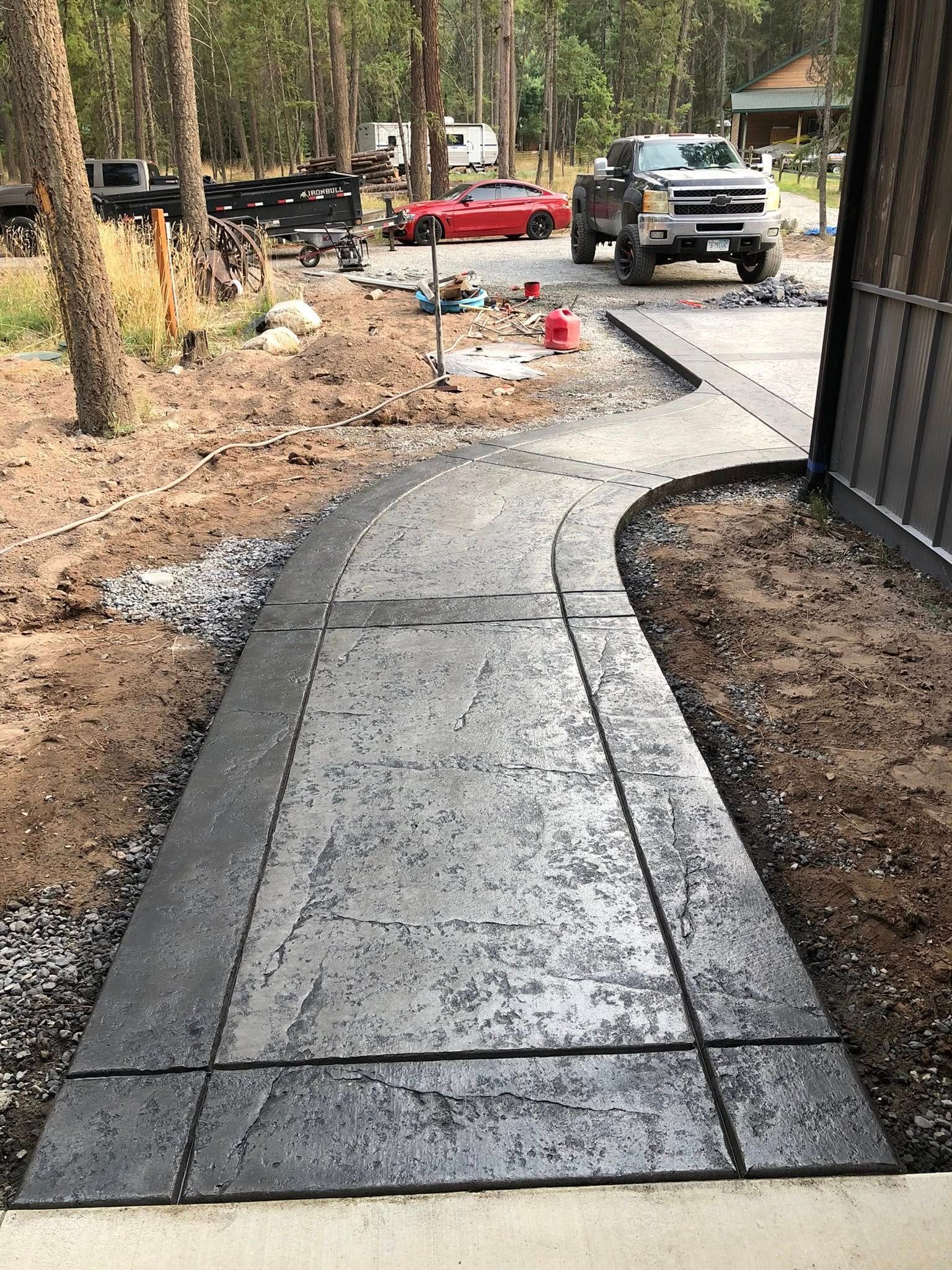Curved concrete walkway under construction beside a driveway with a truck and red car in a wooded area