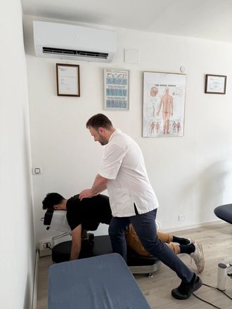 Man helping another on a vibrating platform against a wall. Medical setting, white walls, diagrams.
