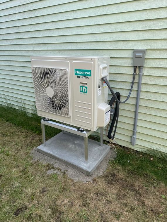 Outdoor heat pump unit mounted on a concrete pad and metal frame, against a light green siding wall.