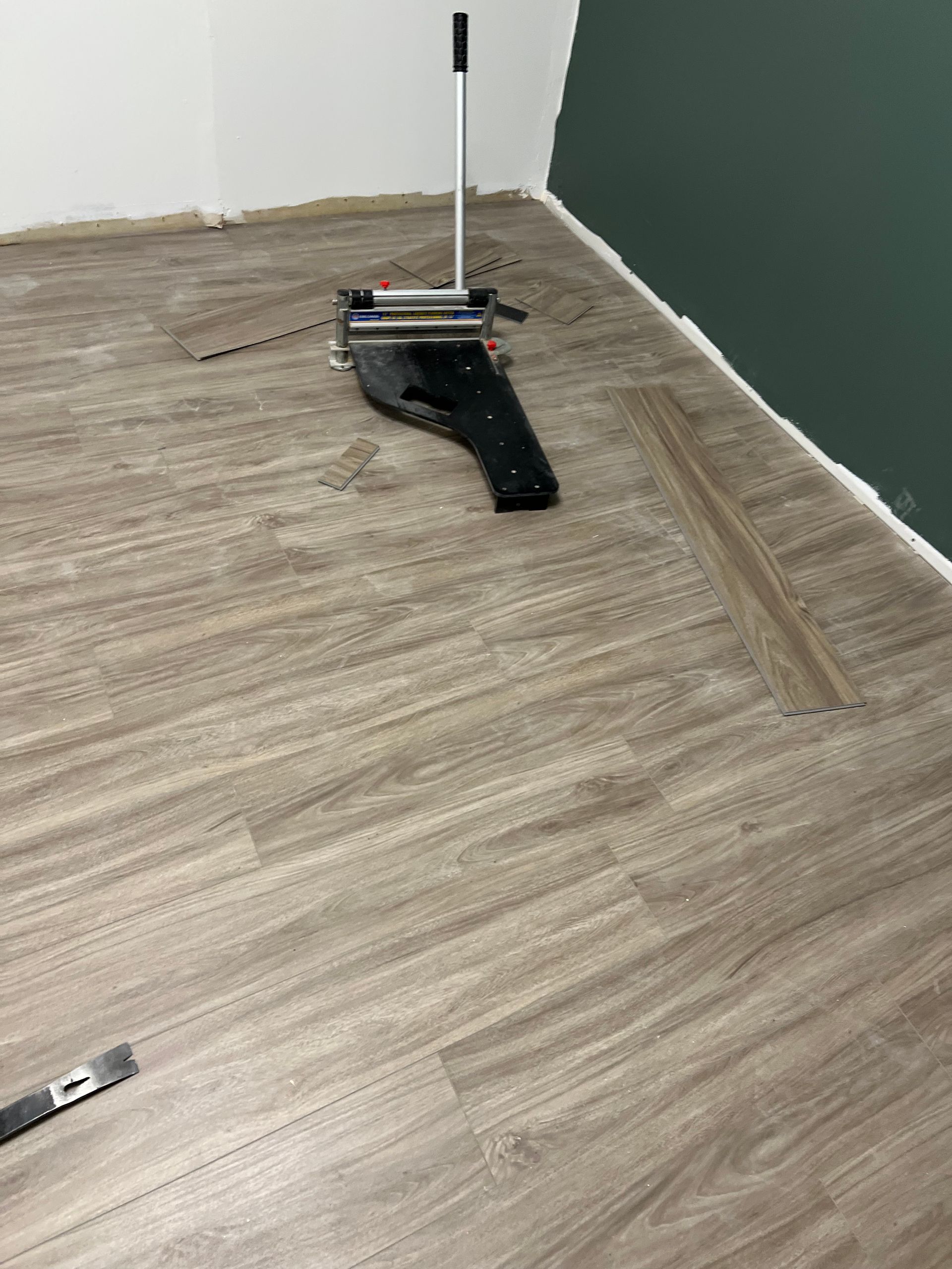 Flooring installation: person using a flooring cutter on light wood-look planks in a room with green and white walls.