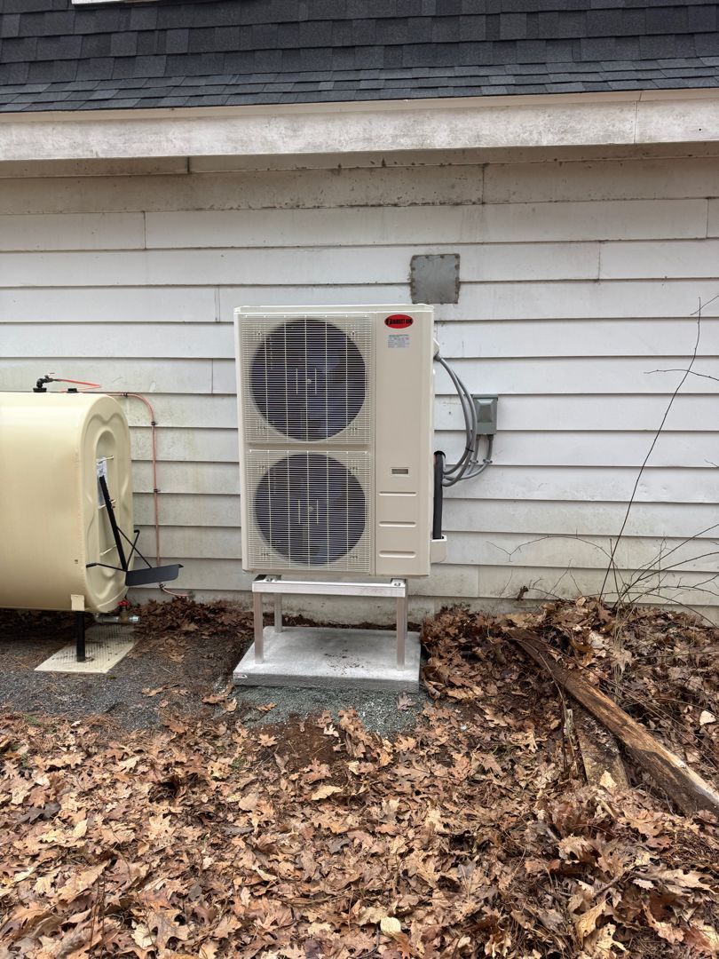 An outdoor HVAC unit and oil tank sit on a gravel base next to a white house, surrounded by dead leaves.