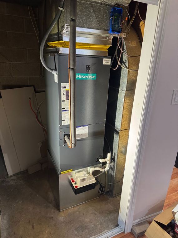 Furnace in a basement, gray metal unit, white pipes and wires, concrete floor.