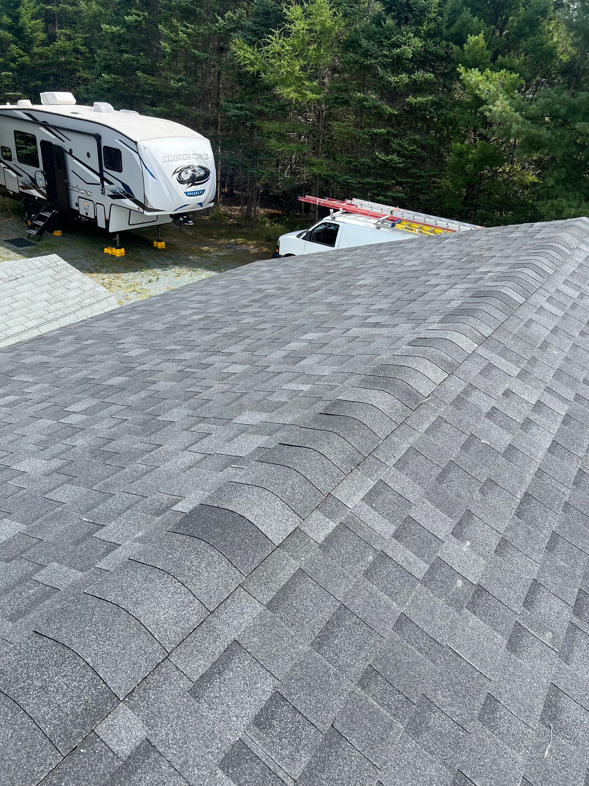Gray asphalt shingle roof, trailer and truck in the background, trees on a sunny day.