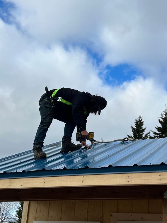 Person on a roof installing metal roofing, wearing a harness, with a cloudy sky in the background.
