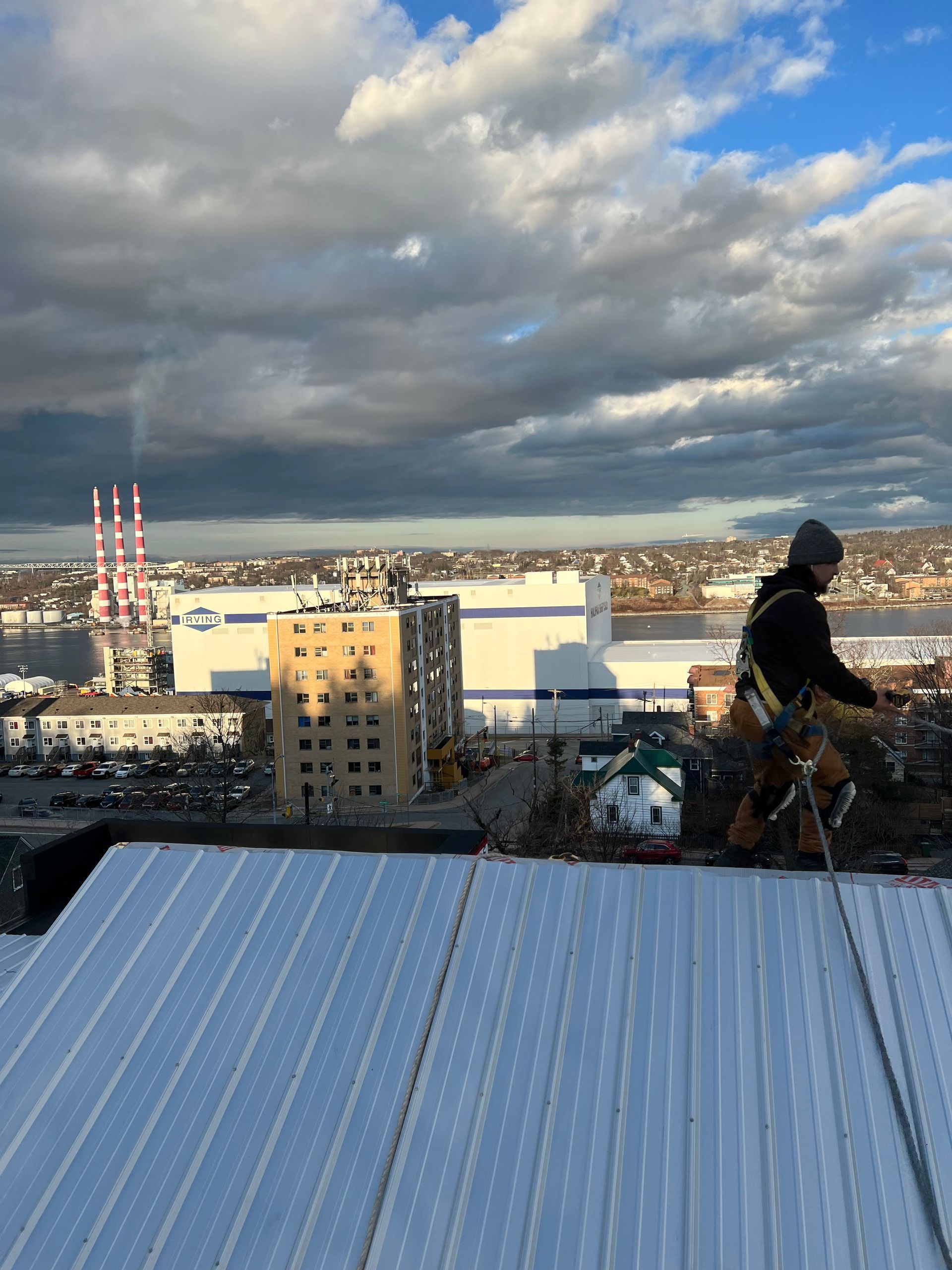 Rooftop worker in harness, installing solar panels against cloudy sky, industrial background with smokestacks and water.