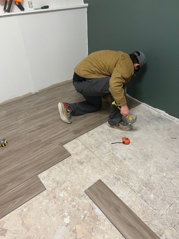 Man installing wood-look flooring on a concrete subfloor in a room with green and white walls.
