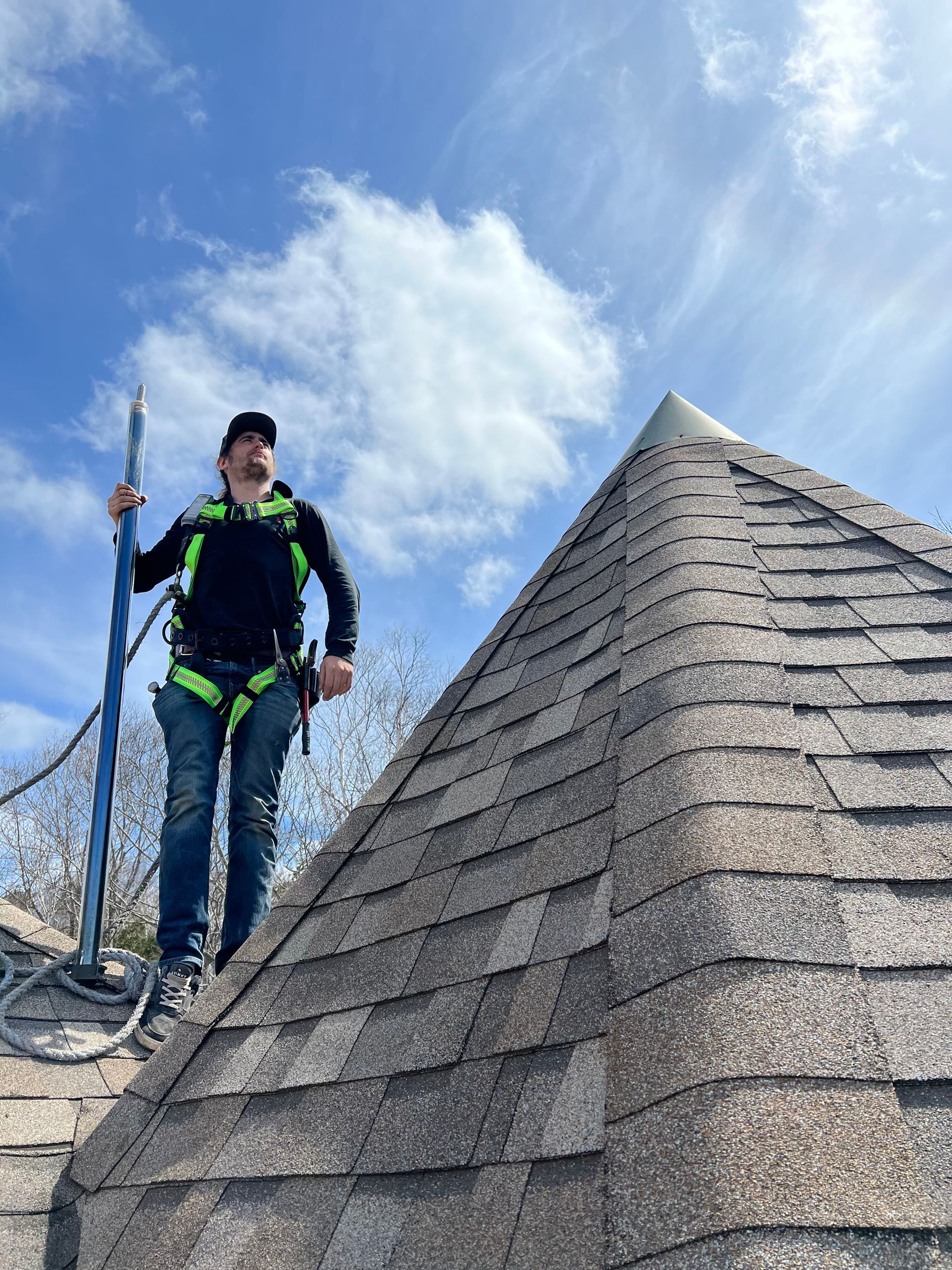Man on pyramid roof with safety harness, holding tool. Blue sky with clouds.