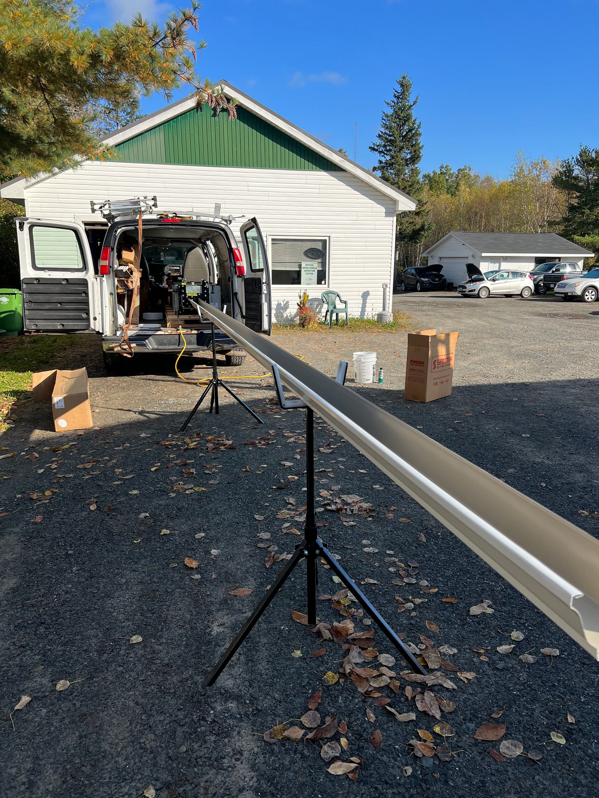 Gutter installation scene: van with open doors, long gutter on stands, gravel driveway, white building.