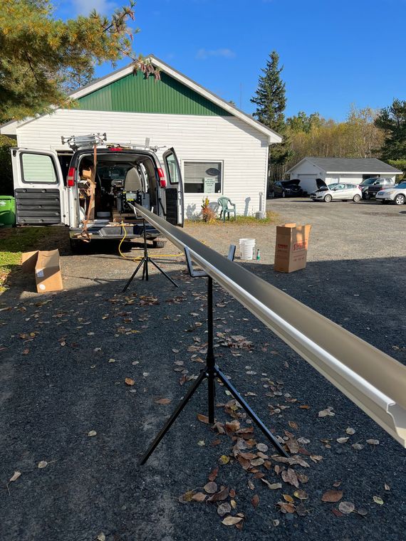 A gutter installation setup: A white van parked near a house, holding a long white gutter on a tripod.
