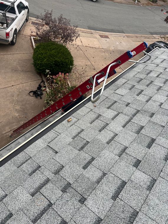 A red ladder with a roof hook is leaning against a gray shingle roof. A white truck is parked nearby.