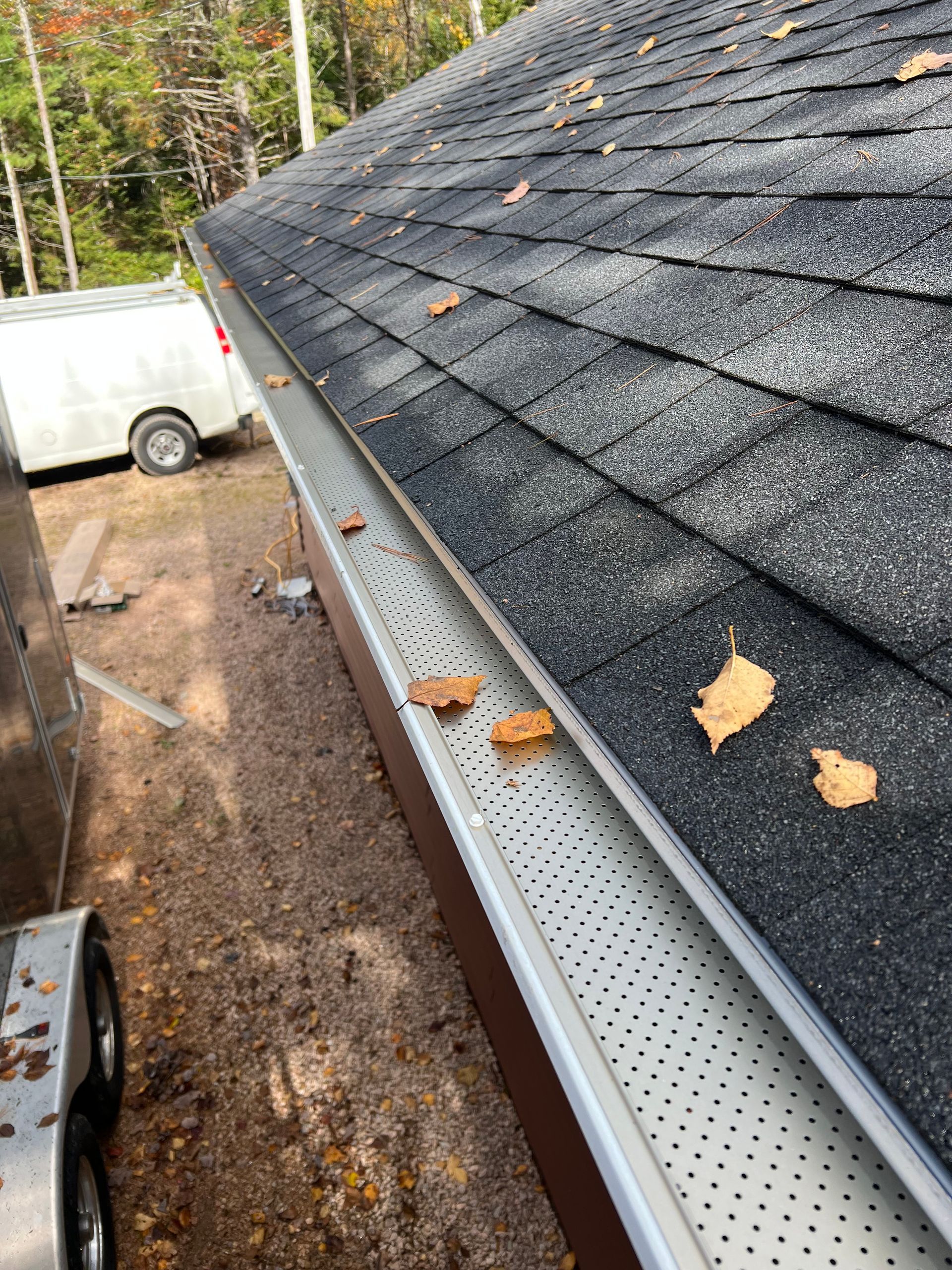 Gutter guard on a shingled roof with leaves, brown trim, a white van, and trailer parked.