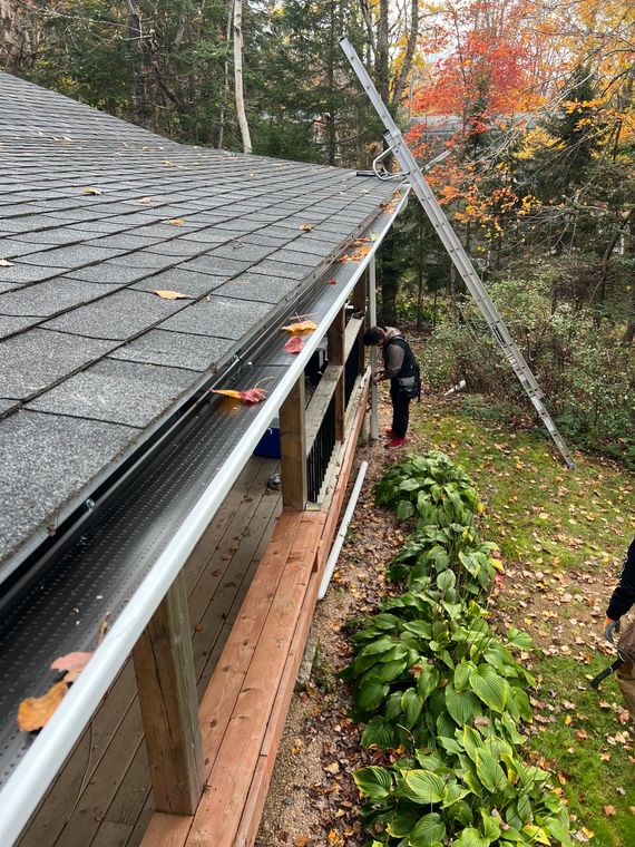 Person cleaning gutters of a house, ladder propped against the roof. Fall foliage visible.