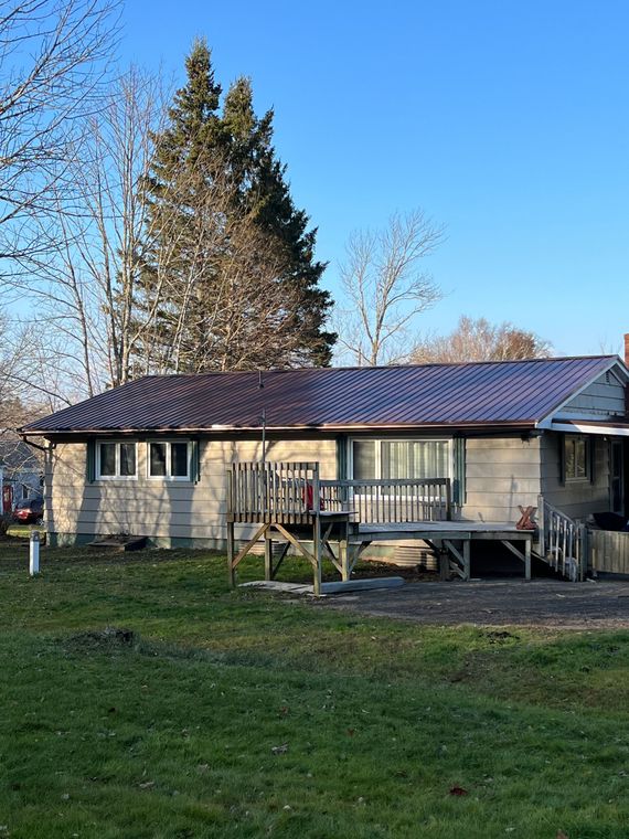 Tan house with a deck, brown roof, and tall trees in the background under a blue sky.