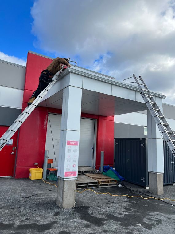 Man on a ladder working on a building's roof edge. Red and white building, blue sky.