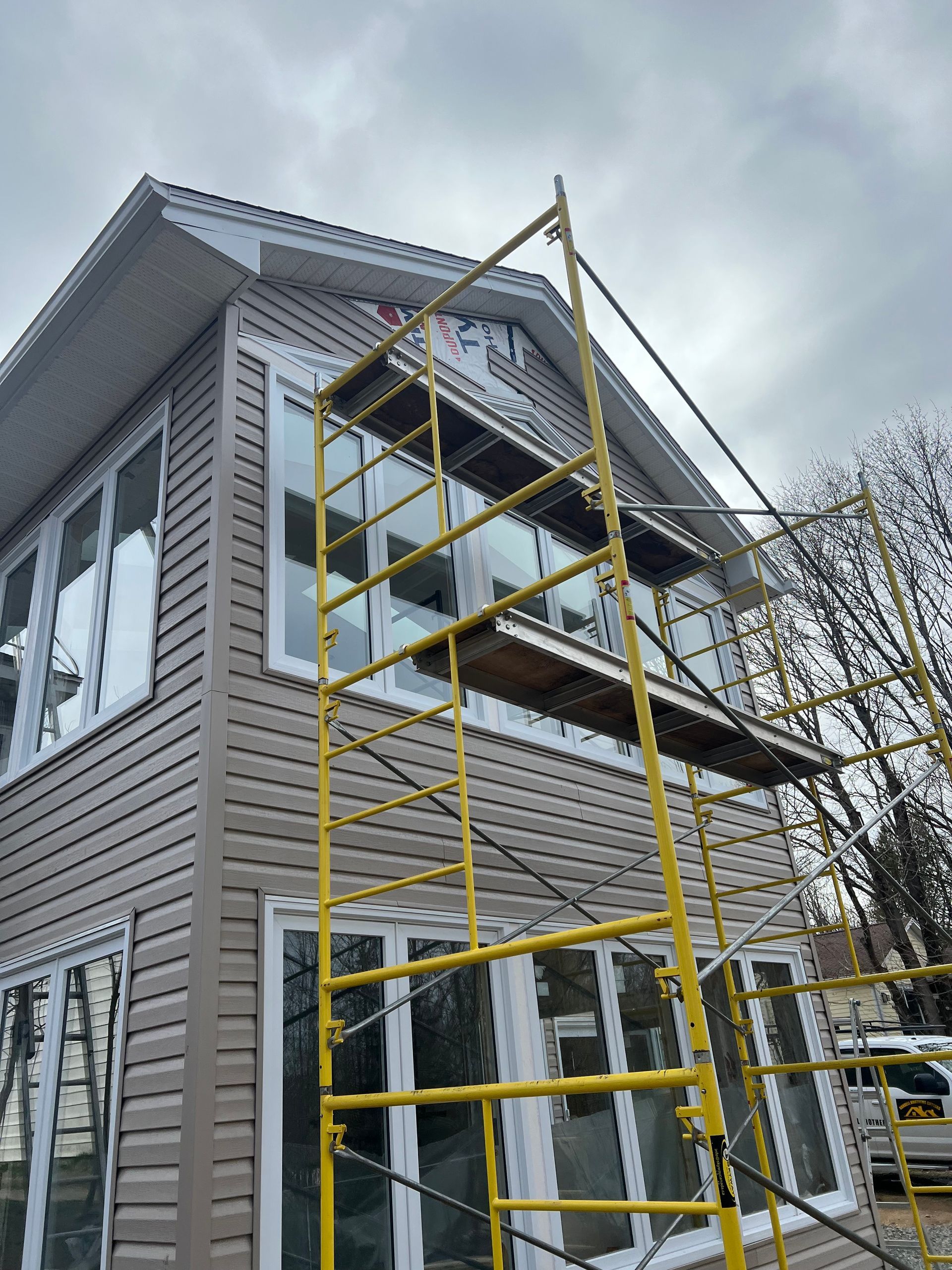 Yellow scaffolding on a house with tan siding and white-framed windows; cloudy sky.