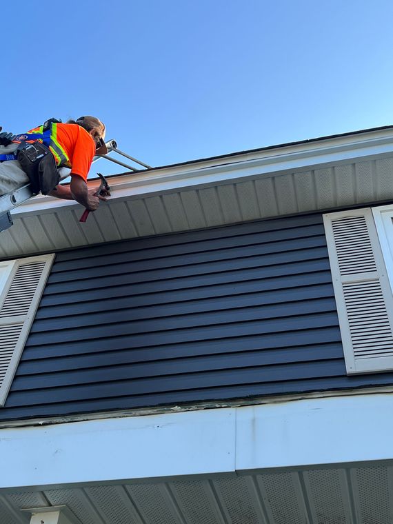 Man in harness working on a house's trim. Blue siding, white shutters, bright orange shirt. Clear blue sky.