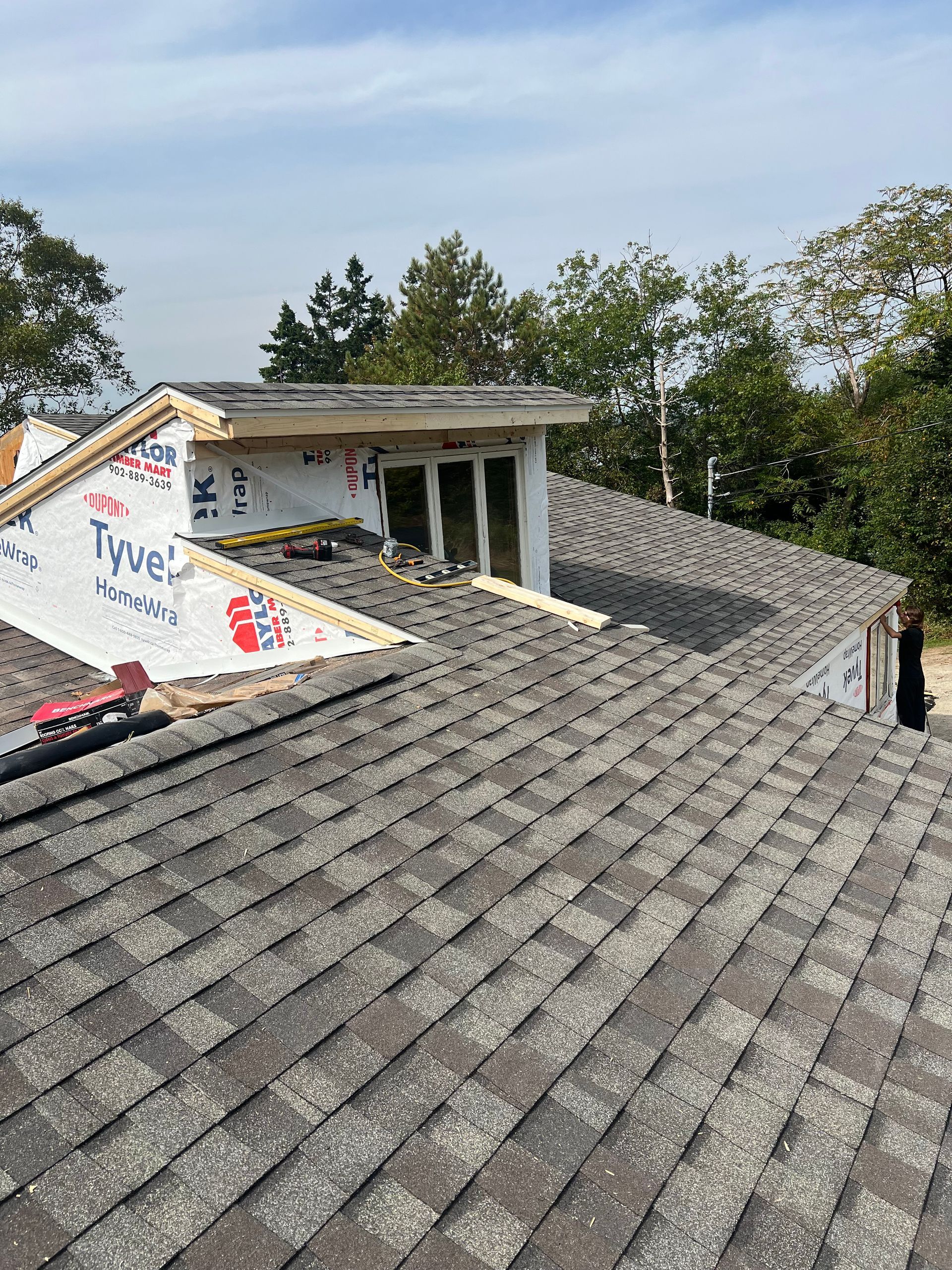 Rooftop with gray shingles, a dormer, and building materials; blue sky and trees in the background.