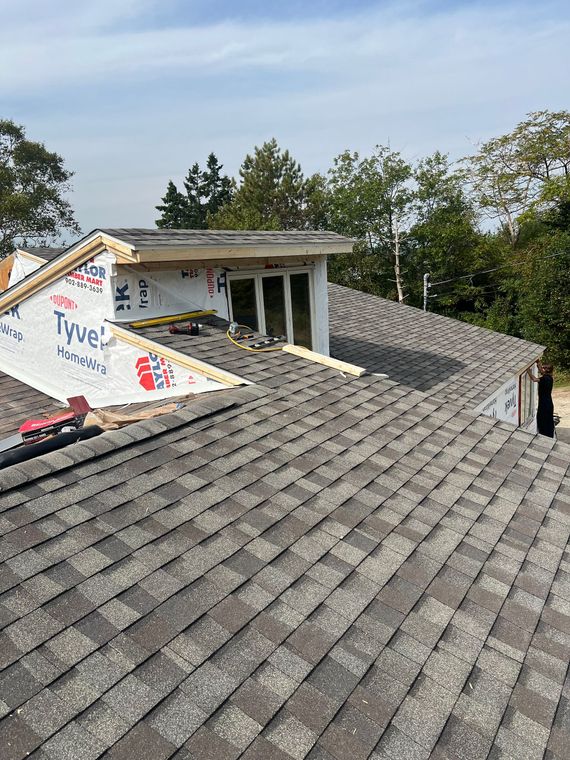 Roofing work in progress, gray shingles, Tyvek wrap, small window structure, trees in background.