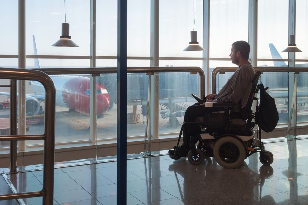 Traveler in wheelchair waiting at an airport terminal window, with a plane visible outside.
