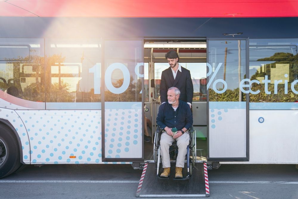 Person in wheelchair on bus ramp, with another person behind, beside a white-and-blue transit bus.