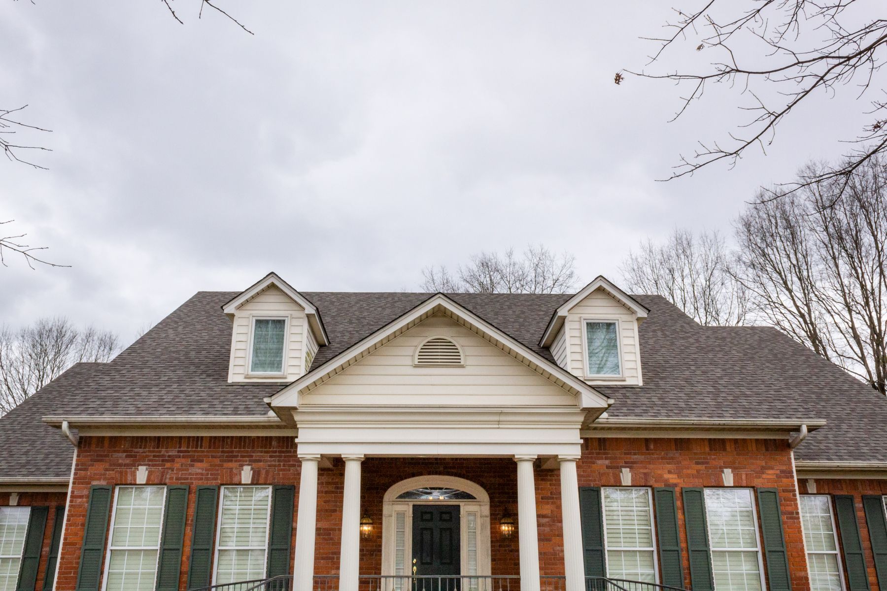 A large brick house with a gray roof and green shutters on a cloudy day.