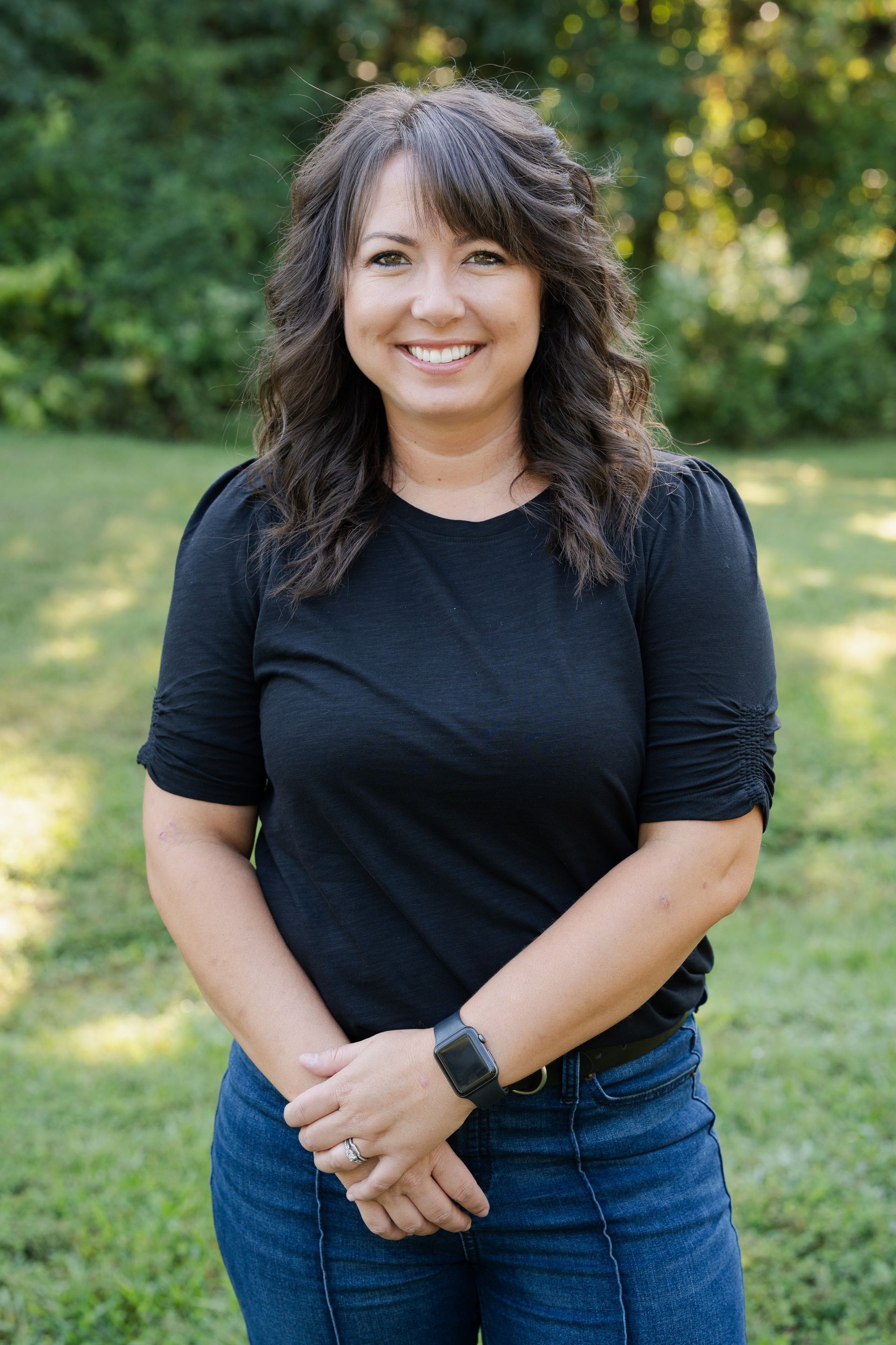 A woman in a black shirt and blue jeans is standing in a field with her hands folded.