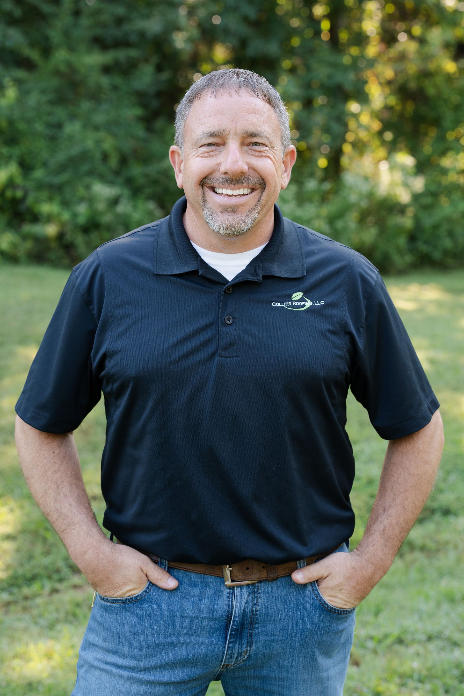 A man in a black shirt and blue jeans is standing in a field with his hands in his pockets.