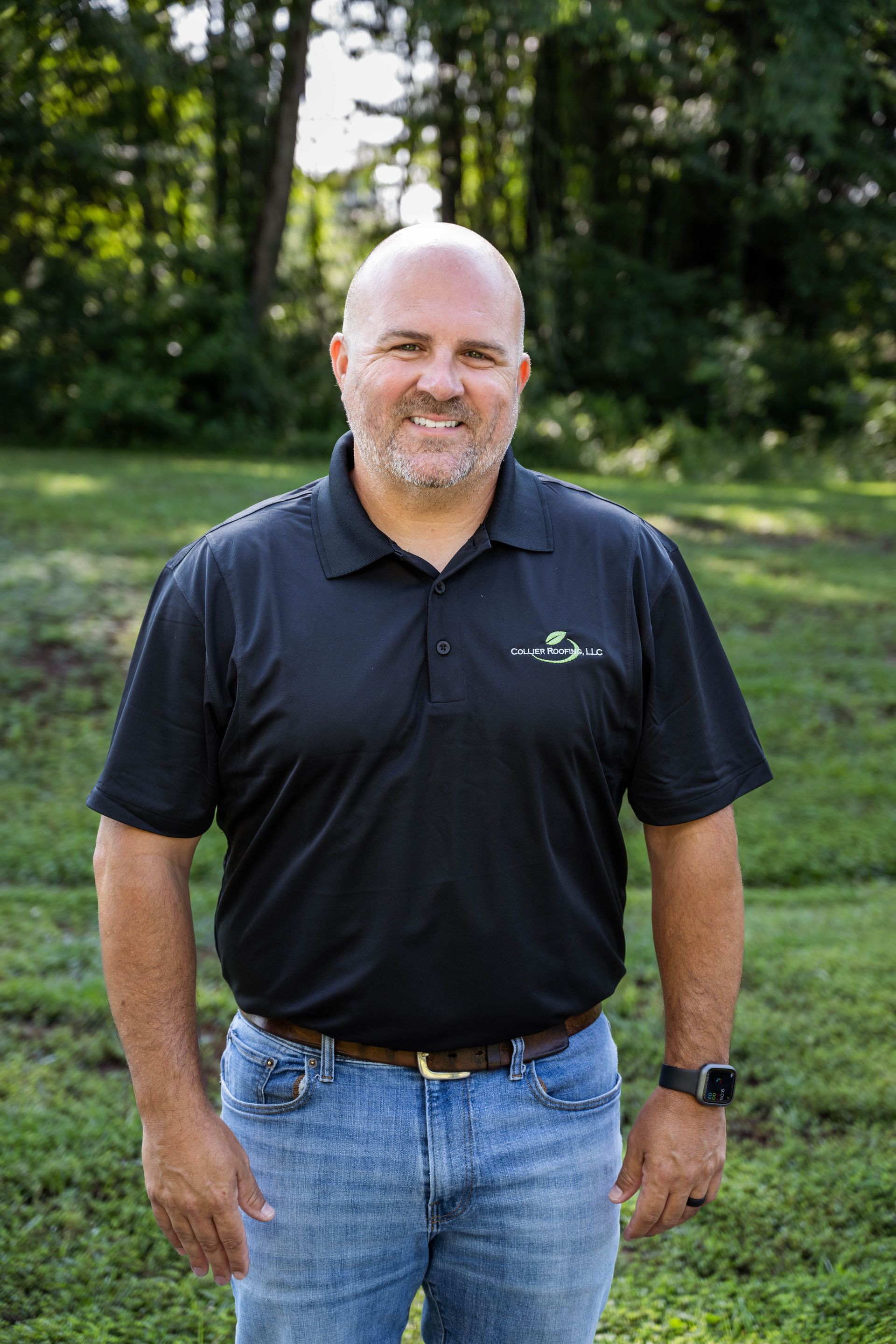 A man in a black shirt and blue jeans is standing in a grassy field.