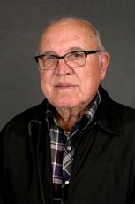 A person wearing glasses, a plaid shirt, and a black jacket, centered against a plain gray background.