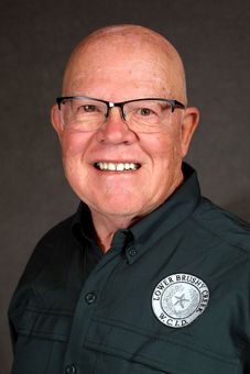 Headshot of a smiling person wearing glasses and a dark green uniform shirt with a circular embroidered patch.