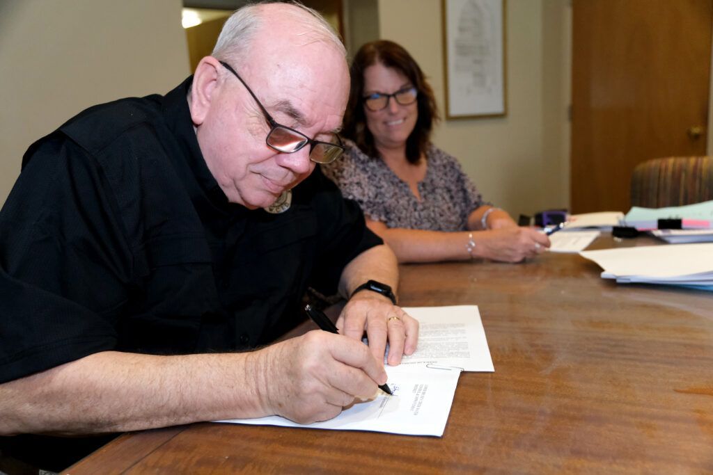 A person in a black shirt signs a document at a wooden table while another person smiles in the background.