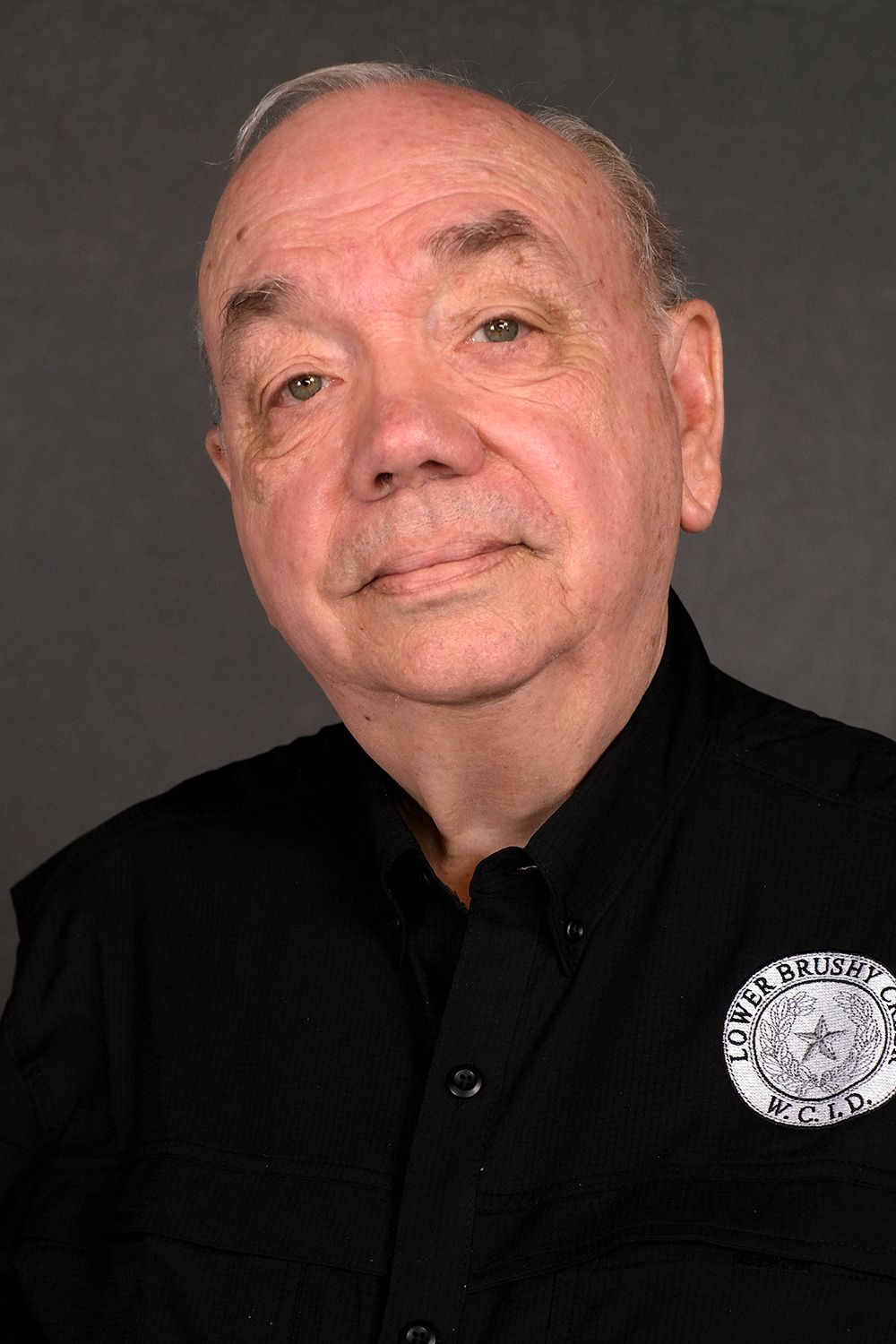 A head-and-shoulders portrait of a person in a black button-down shirt with a circular patch against a dark grey background.