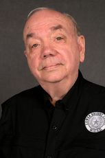 A head-and-shoulders portrait of a person in a black button-down shirt with a circular patch against a dark grey background.
