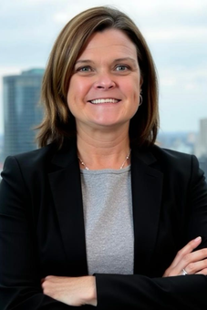 A smiling professional with shoulder-length brown hair, wearing a black blazer and gray shirt, arms crossed against a city.