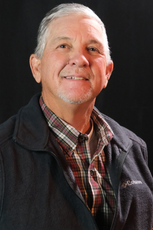 A smiling person wearing a plaid button-down shirt and a dark fleece vest against a black background.