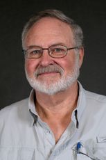 A person with a beard and glasses wearing a light-colored button-down shirt, centered against a dark grey background.