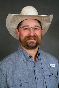 A person with a beard and glasses wears a light-colored straw cowboy hat and a blue button-down shirt against a gray wall.