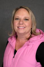 A smiling person with blonde shoulder-length hair wearing a pink hooded vest over a black shirt against a gray background.