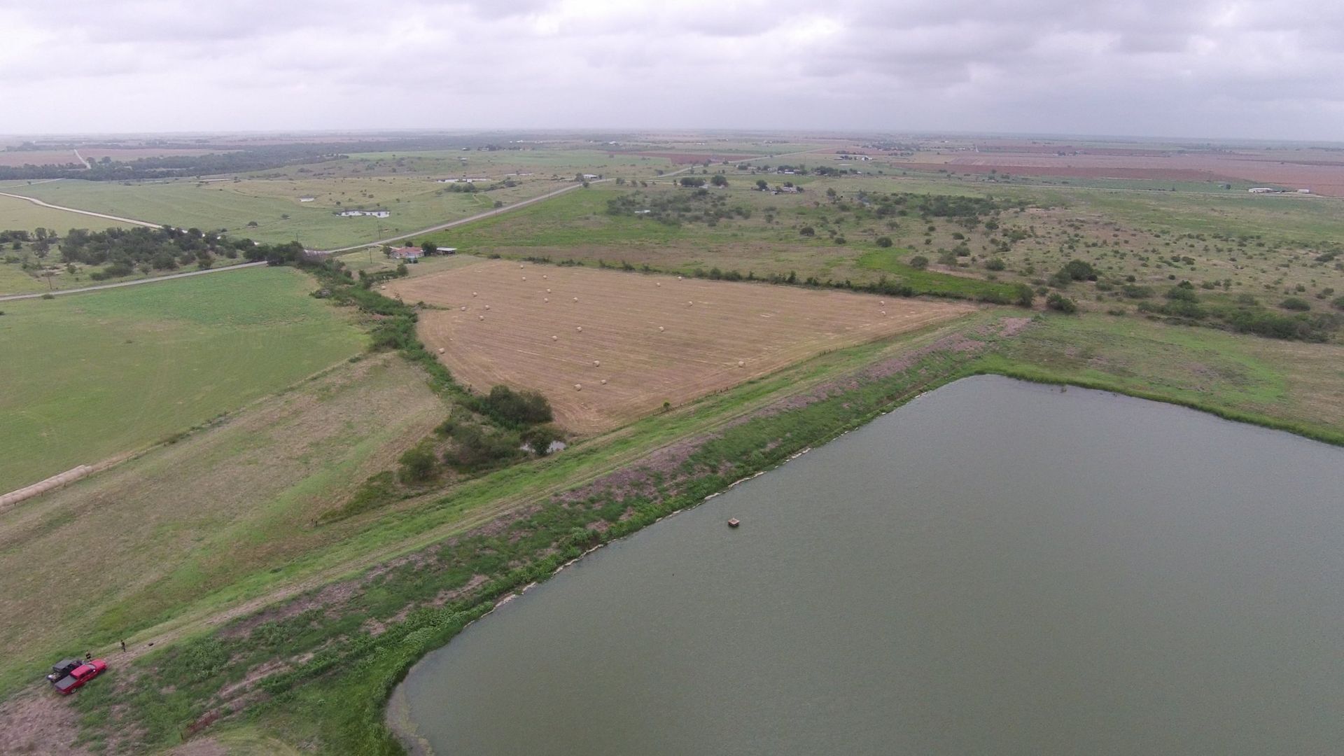 Aerial view of a rectangular pond next to a field of harvested crops, green pasture, and trees under a cloudy sky.
