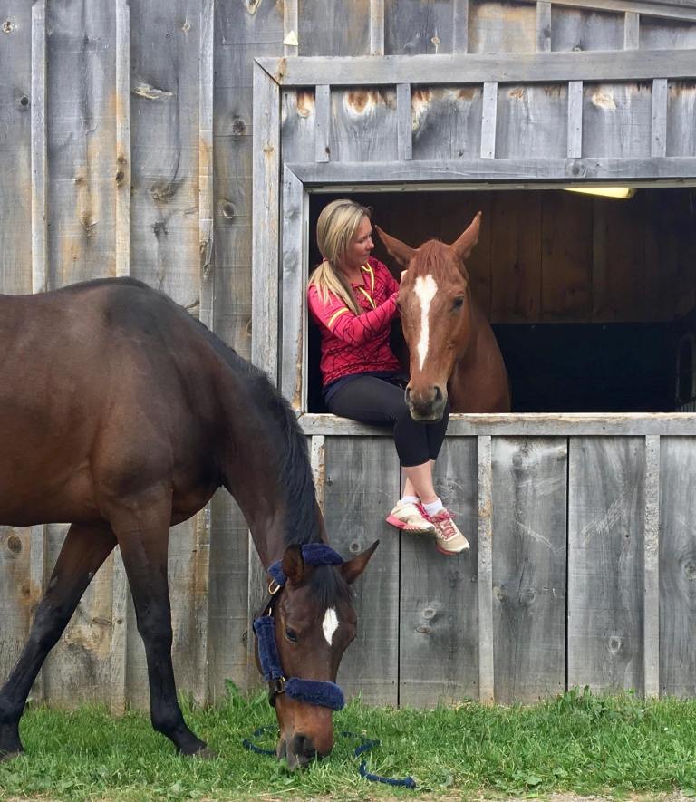 A woman sitting in a barn window combing a horses mane while another horse eats grass outside the barn