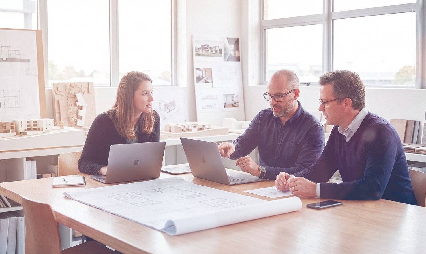 Drei Kollegen arbeiten in einem hellen Büro gemeinsam an einem Tisch mit Laptops und Architekturplänen.