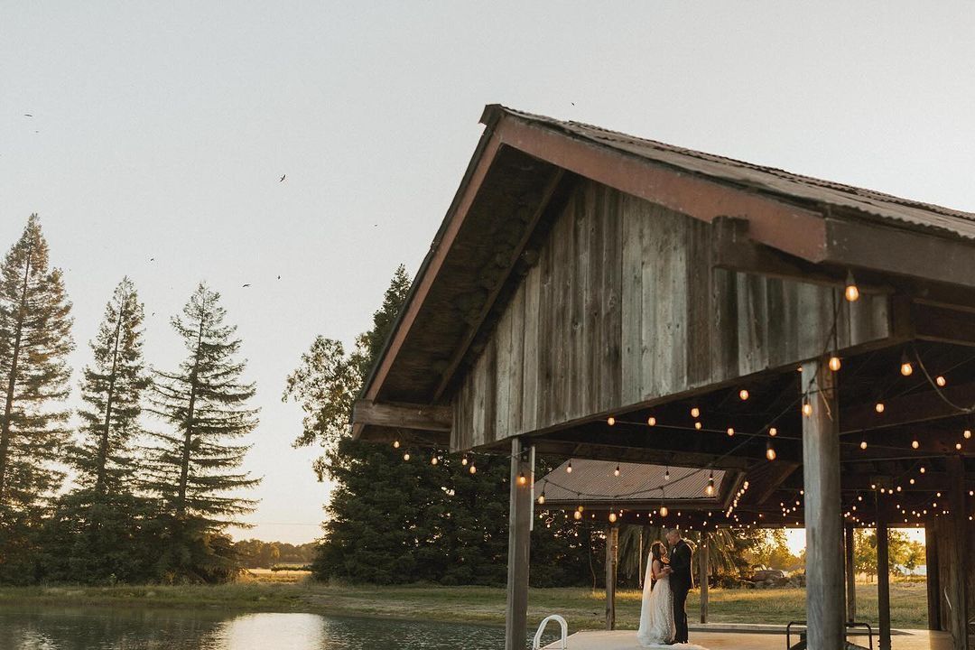 A bride and groom are standing on a dock under a pavilion overlooking a lake.