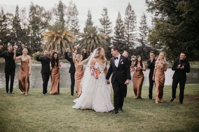 newly married man and woman smile as they hold hands and look at each other with bride's maids and groomsmen cheering in the background on Lakeview Estate's rental space
