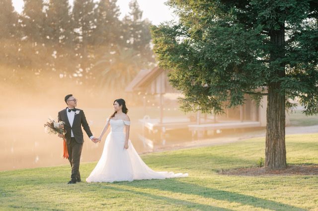 groom leading bride by the hand on a lawn holding a large bouquet, they are mid stride smiling at each other in the daytime with a lake in the background and a dock faded in the background as well