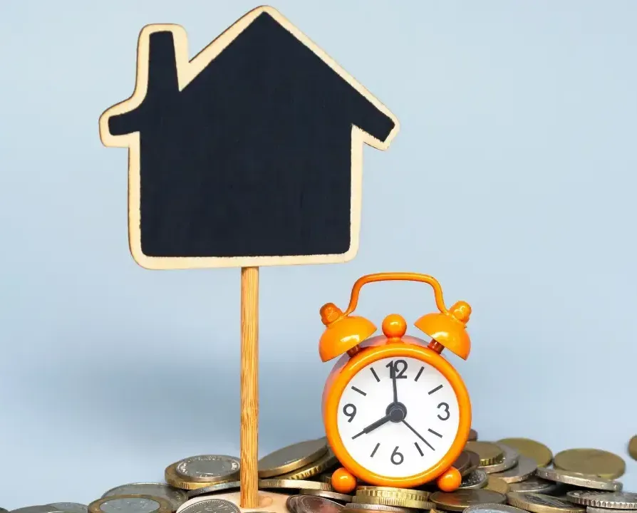 An alarm clock is sitting on top of a pile of coins next to a sign in the shape of a house.