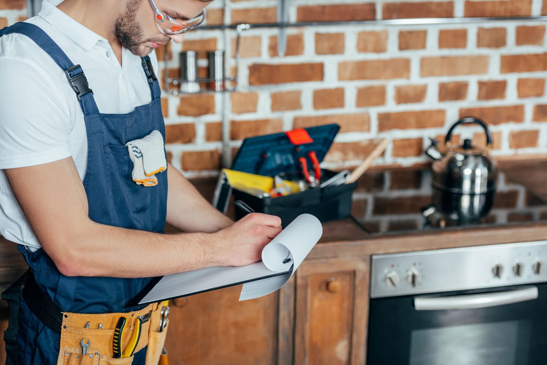 Handyman in overalls and safety glasses writing on a clipboard in a kitchen with tools.