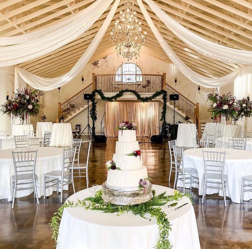 Wedding reception: White cake on a table, round tables with white cloths, draped ceiling, flowers, and chandelier.