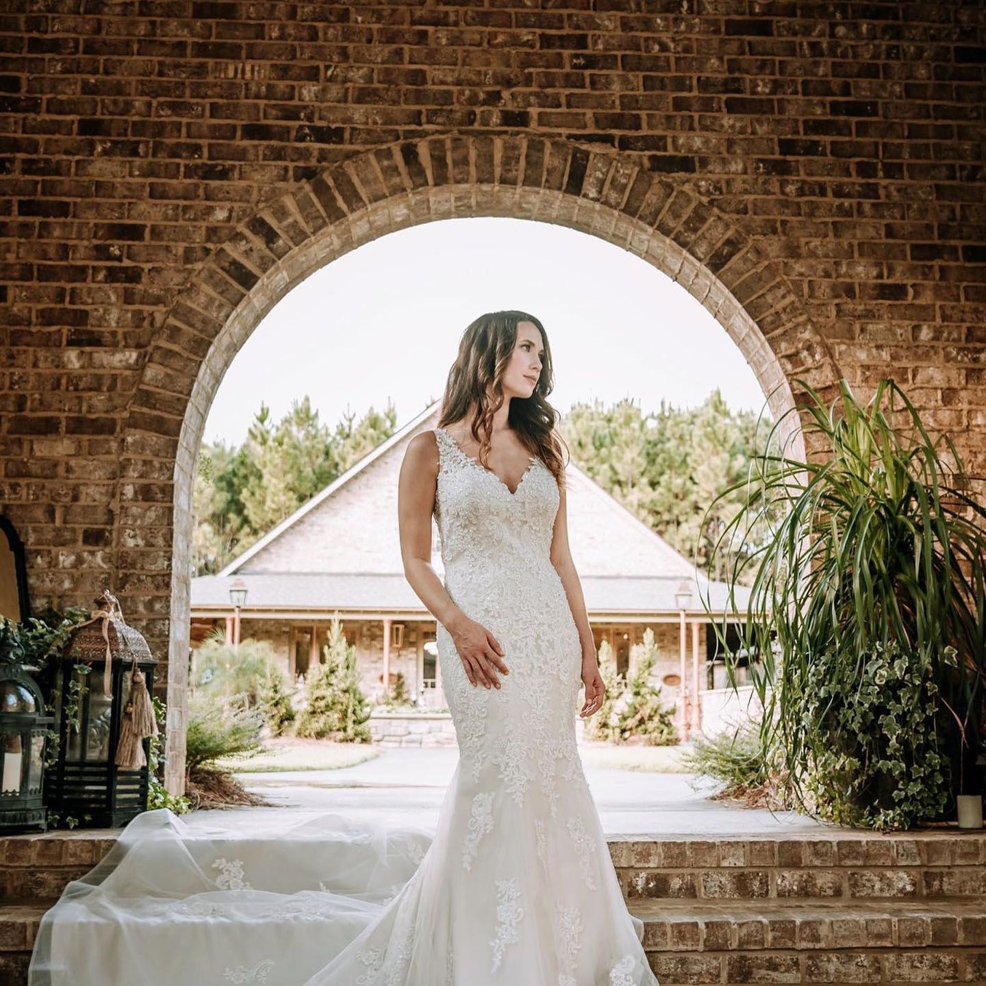 Bride in a lace wedding dress, standing under a brick archway; house and trees in background.